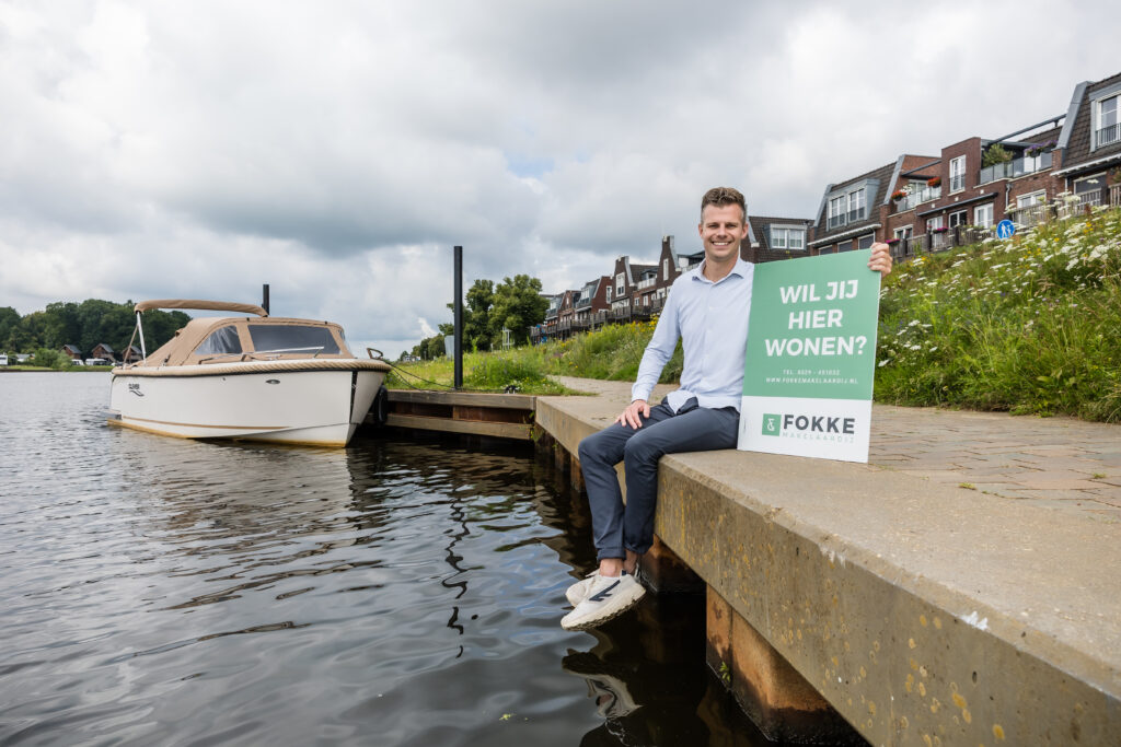 Makelaar zit op een kade aan het water met een bord met de tekst Wil jij hier wonen van Fokke