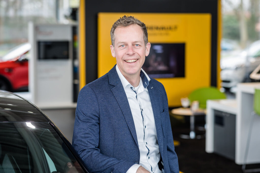 Lachende man in blauw colbert leunt tegen een auto in een autoshowroom